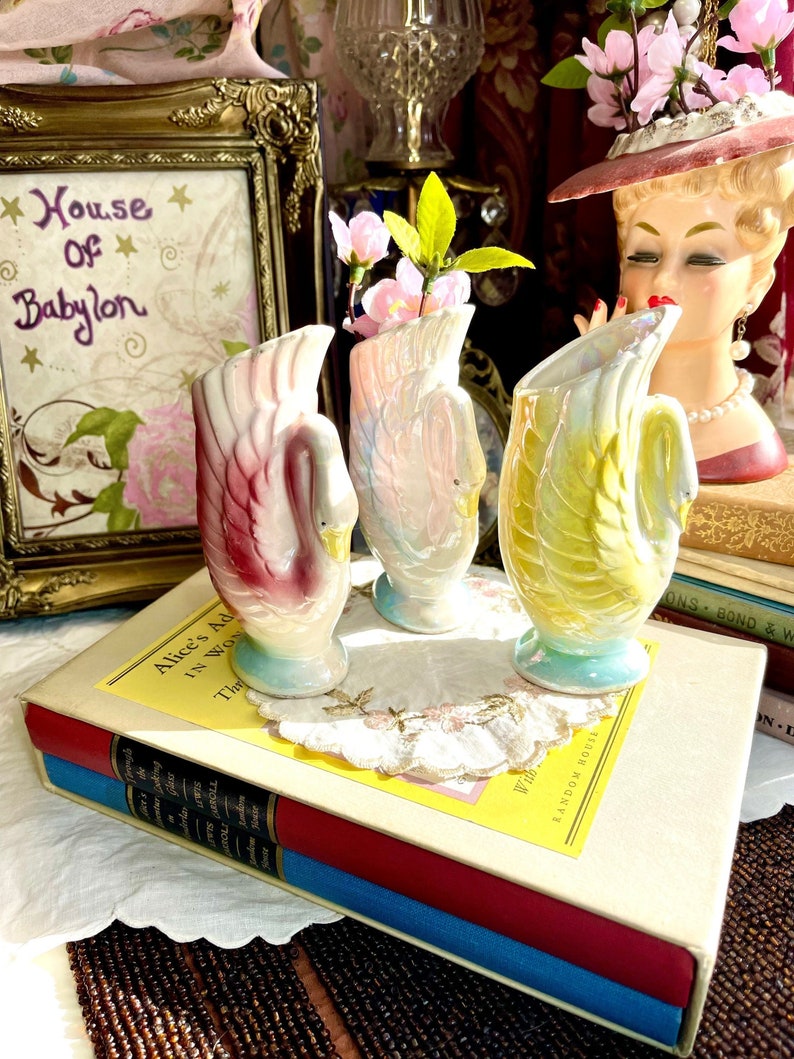 May include: Three iridescent swan-shaped ceramic vases with pink, blue, and yellow accents. The vases are arranged on a book with a white doily and a framed picture with the text "House of Babylon" in the background.