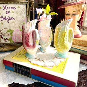 May include: Three swan-shaped ceramic vases with iridescent glaze. The vases are decorated with pink, yellow, and blue colours. The vases are arranged on a white surface with a book and a floral arrangement.