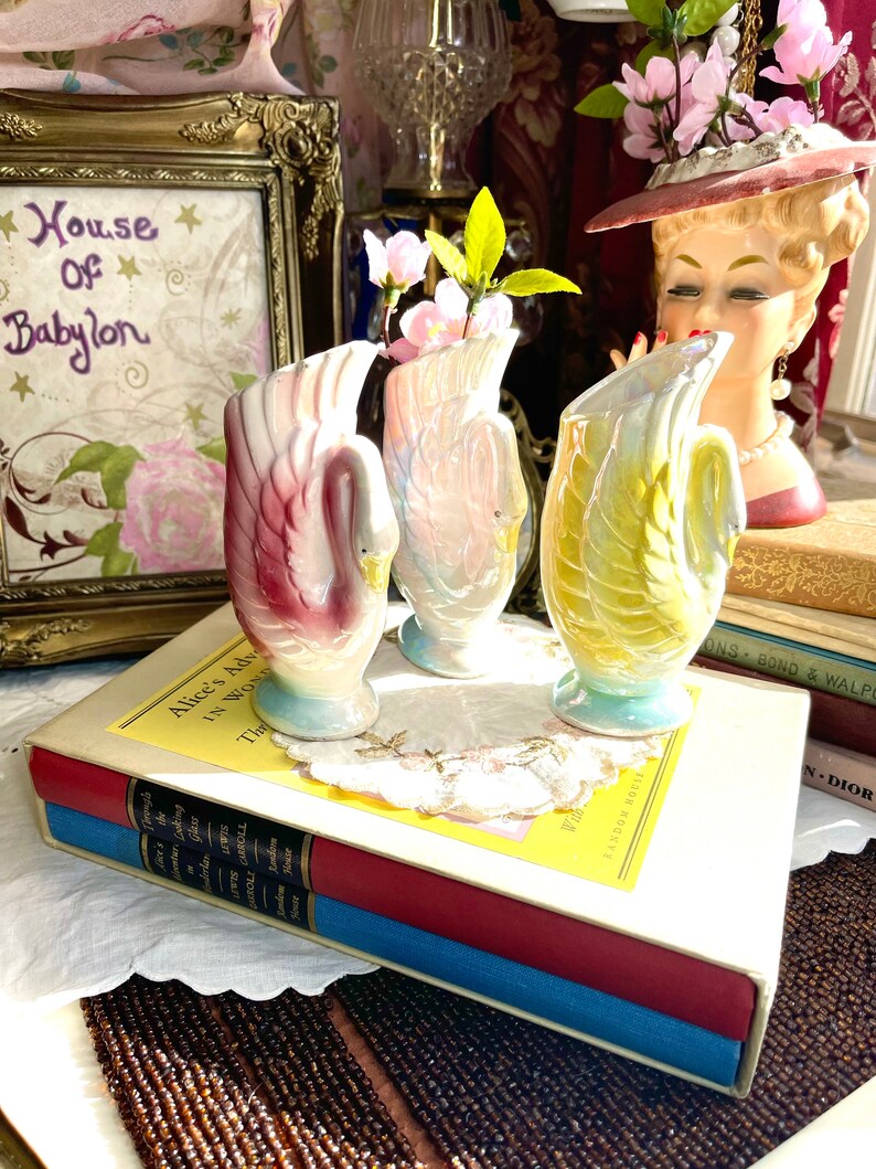 May include: Three swan-shaped ceramic vases with iridescent glaze. The vases are decorated with pink, yellow, and blue colours. The vases are arranged on a white surface with a book and a floral arrangement.