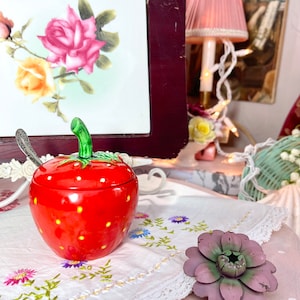 May include: A red and white ceramic strawberry-shaped sugar bowl with a green stem and leaves. The bowl is sitting on a white embroidered tablecloth with a lace trim. A silver spoon is next to the bowl.