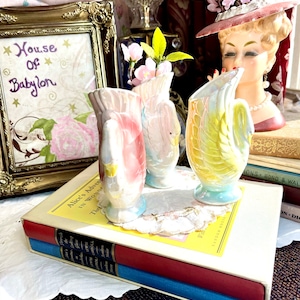 May include: Three vintage ceramic swan-shaped vases with a pink, white and yellow glaze. The vases are arranged on a stack of books with a white cover and a yellow label that reads "Alice's Adventures in Wonderland".