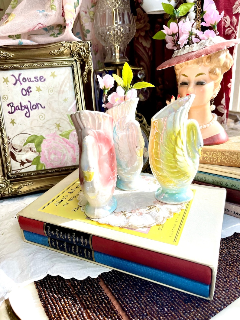 May include: Three vintage ceramic swan-shaped vases with a pink, white and yellow glaze. The vases are arranged on a stack of books with a white cover and a yellow label that reads "Alice's Adventures in Wonderland".