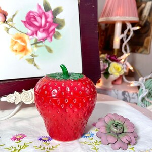 May include: A red glass jar shaped like a strawberry, with a green stem and leaves. The jar is on a white tablecloth with embroidered flowers.