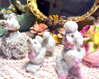 May include: Three white porcelain poodle figurines with pink bows. The poodles are sitting on a white crocheted doily.