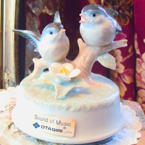May include: A ceramic music box with two blue and white birds on a branch with a flower. The white base has a light blue accent and a label that reads "Sound of Music OTIGIRI JAPAN".