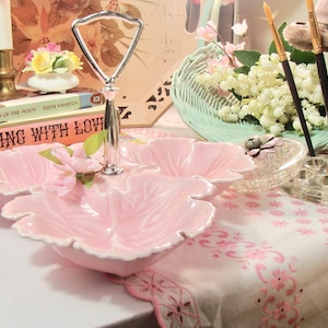 May include: A pink three-section serving dish with a silver handle. The dish is shaped like a flower and has a white rim. The dish is sitting on a white tablecloth with a pink floral pattern. The text "Cooking with Love" is visible on a book in the background.