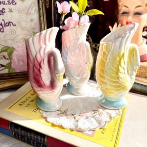 May include: Three iridescent swan-shaped ceramic vases with pastel pink, blue, and yellow glaze. The vases are arranged on a white doily with a book in the background.