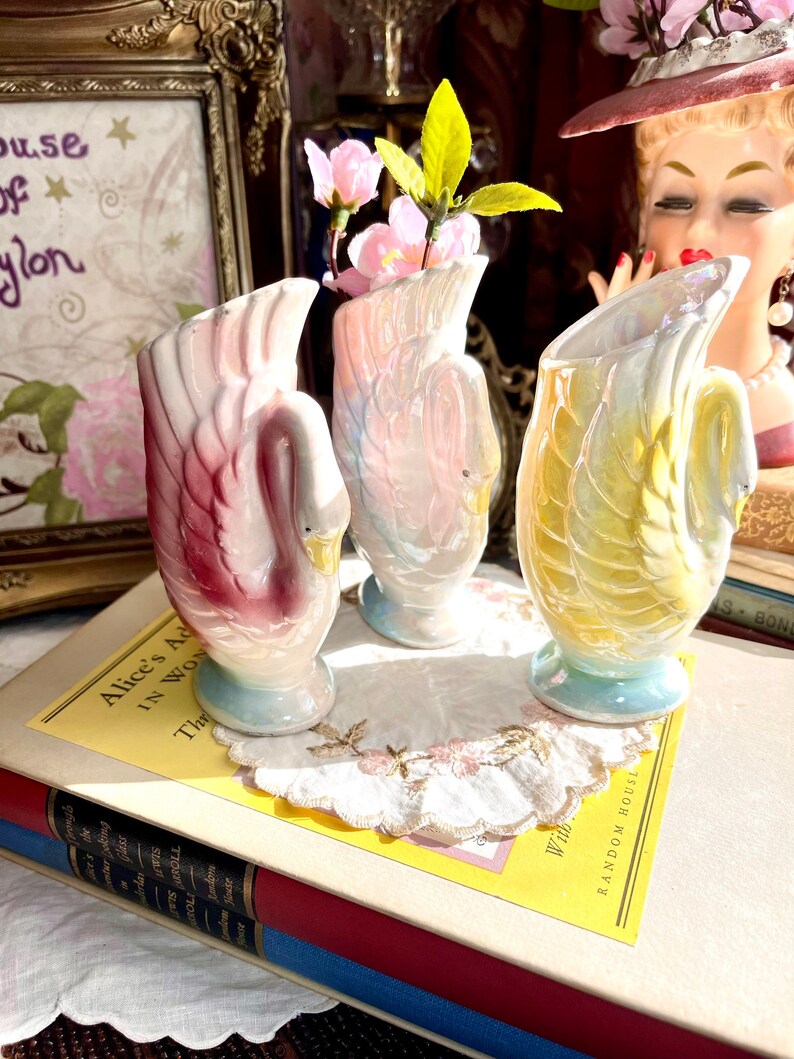May include: Three iridescent swan-shaped ceramic vases with pastel pink, blue, and yellow glaze. The vases are arranged on a white doily with a book in the background.