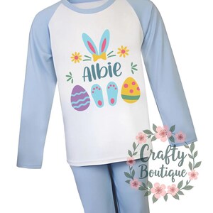 以下が含まれることがあります： 青い長袖のパジャマに、ウサギの耳のデザイン、名前の「Albie」とイースターエッグが描かれています。