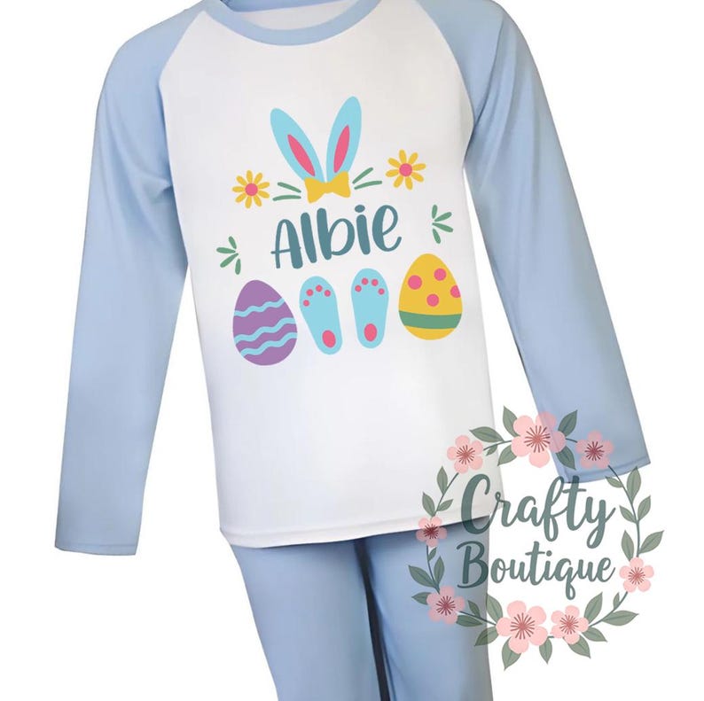 以下が含まれることがあります： 青い長袖のパジャマに、ウサギの耳のデザイン、名前の「Albie」とイースターエッグが描かれています。