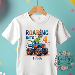 May include: White t-shirt with a graphic of a green dinosaur wearing sunglasses and a party hat, riding a blue monster truck. The text "Roaring into 4" is above the truck and the name "Finley" is below the truck.