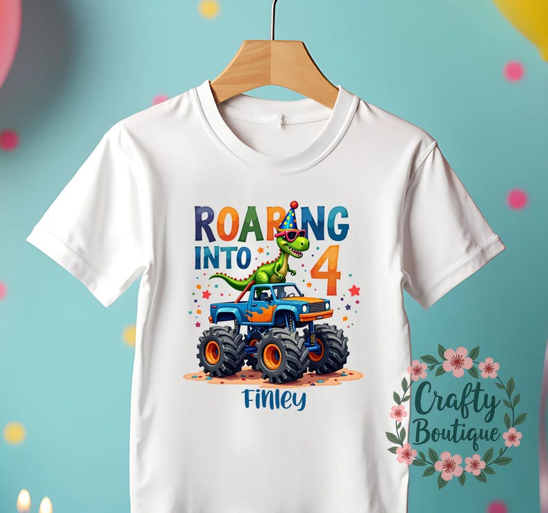 May include: White t-shirt with a graphic of a green dinosaur wearing sunglasses and a party hat, riding a blue monster truck. The text "Roaring into 4" is above the truck and the name "Finley" is below the truck.