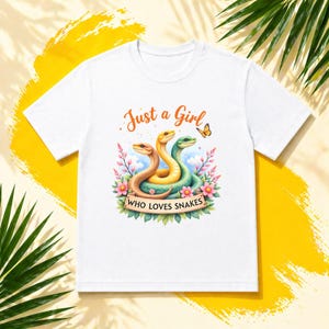 May include: White t-shirt with a colourful illustration of three snakes in brown, yellow, and green, surrounded by flowers and a butterfly. The text "Just a Girl Who Loves Snakes" is displayed above and below the snakes.