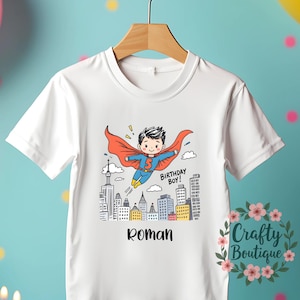 May include: White t-shirt with a cartoon superhero boy design. The boy has a red cape and is flying over a city skyline. The text "BIRTHDAY BOY!" and the name "Roman" are also on the shirt. The number 5 is on the superhero's chest.