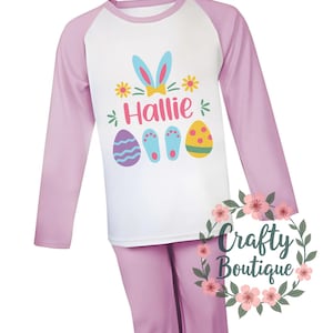 以下が含まれることがあります： ピンクと白の長袖パジャマ。白いラグランスリーブのシャツには、リボンをつけたウサギ、名前の「Hallie」、イースターエッグが描かれています。