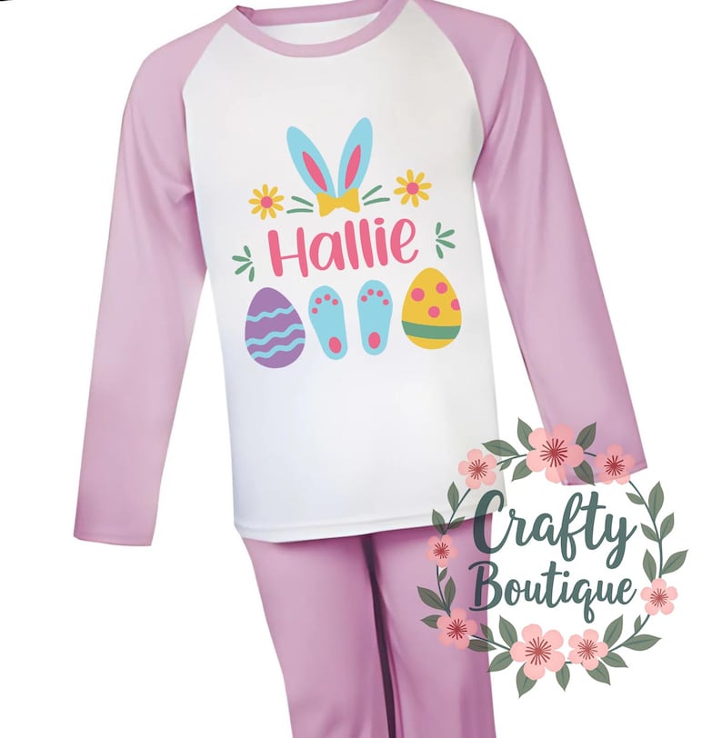 以下が含まれることがあります： ピンクと白の長袖パジャマ。白いラグランスリーブのシャツには、リボンをつけたウサギ、名前の「Hallie」、イースターエッグが描かれています。