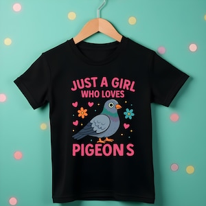 May include: Black t-shirt with the text "JUST A GIRL WHO LOVES PIGEONS" and a cartoon pigeon illustration. The design includes pink hearts and flowers. The shirt is hanging on a wooden hanger.