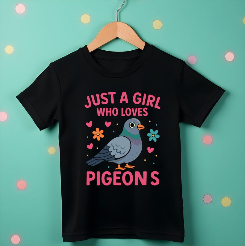 May include: Black t-shirt with the text "JUST A GIRL WHO LOVES PIGEONS" and a cartoon pigeon illustration. The design includes pink hearts and flowers. The shirt is hanging on a wooden hanger.