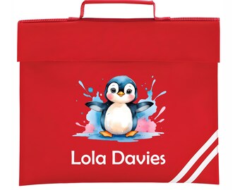 May include: A red school satchel with a handle and a flap closure. The front features a watercolor illustration of a penguin with blue and black wings, standing in front of blue and pink splashes. The name "Lola Davies" is printed in white.