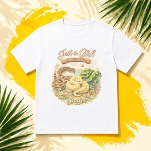May include: White t-shirt with a colourful illustration of three snakes: a brown and tan snake, a yellow snake, and a green snake. The text "Just a Girl Who Loves Snakes" is above the snakes.