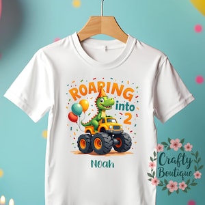 May include: White t-shirt featuring a green dinosaur in a party hat driving a yellow monster truck. The text "Roaring into 2" and the name "Noah" are displayed in a colourful design, suitable for a child's birthday.
