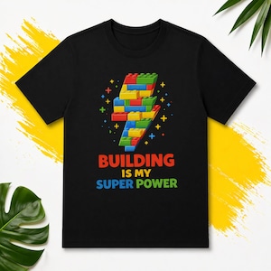 May include: Black t-shirt with a graphic of a lightning bolt constructed from toy bricks. The text "BUILDING IS MY SUPER POWER" is in red, blue, and green. The design includes stars and colourful accents.