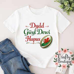 May include: White t-shirt with the text "Dydd Gwyl Dewi Hapus" in red and green script. A rugby ball graphic with the Welsh flag is on the right. The shirt is paired with blue jeans.