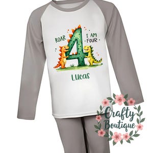 May include: Gray and white long-sleeved pajamas with a dinosaur design. The shirt features a green number four with two dinosaurs on either side. The text "Roar" and "I am Four" is printed on the shirt. The name "Lucas" is printed below the number four.