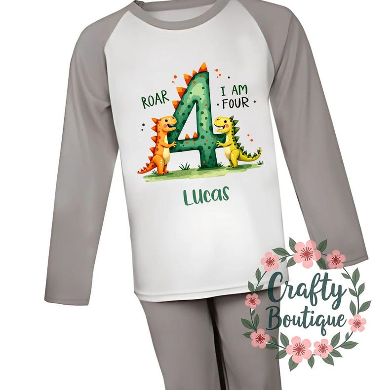 May include: Gray and white long-sleeved pajamas with a dinosaur design. The shirt features a green number four with two dinosaurs on either side. The text "Roar" and "I am Four" is printed on the shirt. The name "Lucas" is printed below the number four.