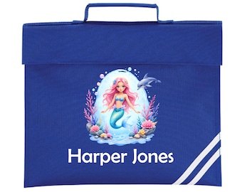 May include: A royal blue school satchel with a handle and flap closure. The front features a mermaid illustration with pink hair and a turquoise tail, a dolphin, and the name "Harper Jones" in white. White stripes accent the lower right corner.