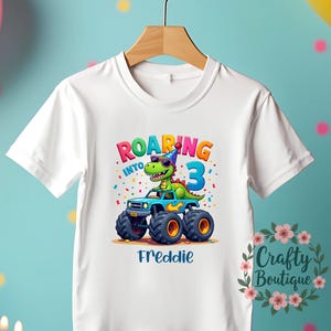 May include: White t-shirt featuring a colourful graphic of a dinosaur wearing a party hat and driving a monster truck. The text "Roaring into 3" is above the graphic and the name "Freddie" is below.