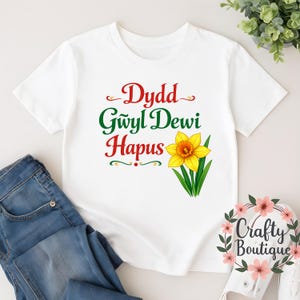 Puede incluir: Camiseta blanca con el texto "Dydd Gwyl Dewi Hapus" en rojo y verde, y una ilustración de narciso amarillo. La camiseta se combina con unos vaqueros azules. Se ve una corona floral con el texto "Crafty Boutique".
