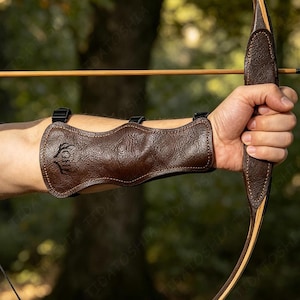 May include: A dark brown leather forearm guard featuring a textured surface, visible stitching along the edges, and three black adjustable buckle straps. The guard displays a small embossed antler logo on the side and is worn while holding a wooden bow.