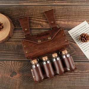 Könnte beinhalten: Ein brauner Leder-Trankflaschenhalter mit vier Glasflaschen und Korken. Der Halter hat eine Klappe mit dem eingravierten Namen "Alexander". Die Flaschen werden in einzelnen Lederhülsen gehalten. Der Halter hat Riemen und Messingnieten.