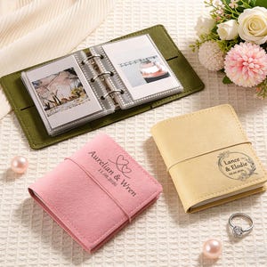 May include: A collection of personalized photo albums in various colors. A green album is open, displaying photos. A pink album is inscribed with "Aurelian & Wren 11.08.2026". A yellow album is inscribed with "Lance & Elodie 08.06.2026". A ring and pearls are also present.