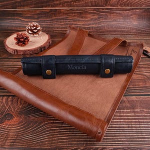 Peut inclure: Un rouleau en cuir bleu avec le mot "Moncia" en relief, reposant sur une surface en cuir marron.