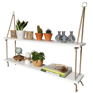 May include: A two-tier white wooden hanging shelf suspended by thick tan jute ropes with metal rings at the top. The shelves hold various items including potted plants, cacti, glass vases, a small globe, a white candle, and stacked books.