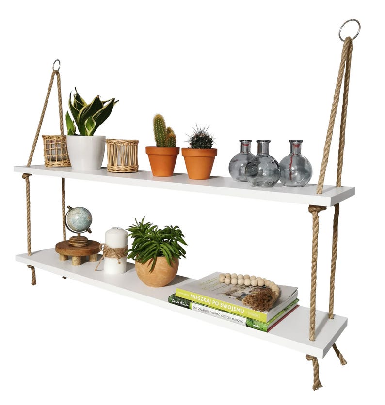 May include: A two-tier white wooden hanging shelf suspended by thick tan jute ropes with metal rings at the top. The shelves hold various items including potted plants, cacti, glass vases, a small globe, a white candle, and stacked books.