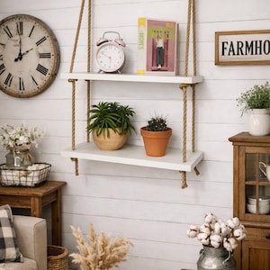 May include: A two-tier wall-mounted shelf featuring two rectangular white wooden boards suspended by thick tan jute ropes. The top shelf holds a pink analog alarm clock and a book, while the bottom shelf displays a green succulent and a cactus.