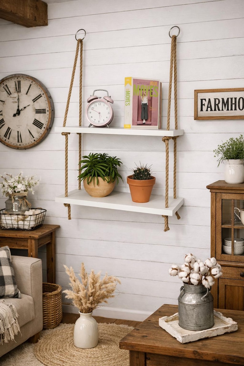 May include: A two-tier wall-mounted shelf featuring two rectangular white wooden boards suspended by thick tan jute ropes. The top shelf holds a pink analog alarm clock and a book, while the bottom shelf displays a green succulent and a cactus.