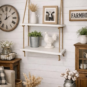 May include: A two-tier white wooden wall shelf suspended by thick tan jute ropes. The top shelf holds a white pitcher with dried grass and a framed cow photograph. The bottom shelf features a galvanized metal bucket with greenery and a white rooster.