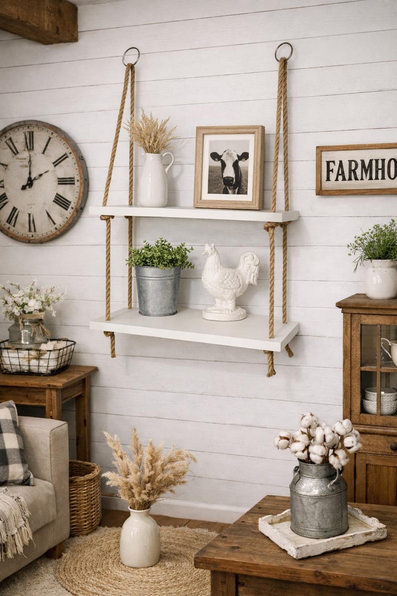 May include: A two-tier white wooden wall shelf suspended by thick tan jute ropes. The top shelf holds a white pitcher with dried grass and a framed cow photograph. The bottom shelf features a galvanized metal bucket with greenery and a white rooster.