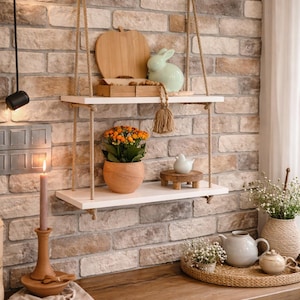 May include: A two-tier white wooden hanging shelf suspended by thick tan rope. The top shelf holds a wooden apple-shaped board and a light green ceramic rabbit. The bottom shelf features a potted orange flowering plant and a small white bird figurine.