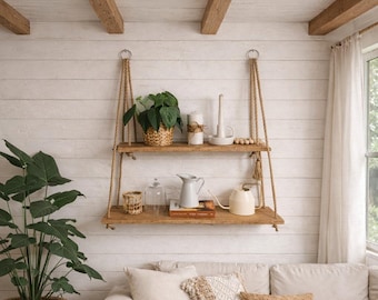 May include: A two-tiered wooden hanging shelf with rope supports. The top shelf holds a potted plant, candles, and decorative beads. The bottom shelf displays a glass cloche, a pitcher, and a watering can. The background is a white shiplap wall.
