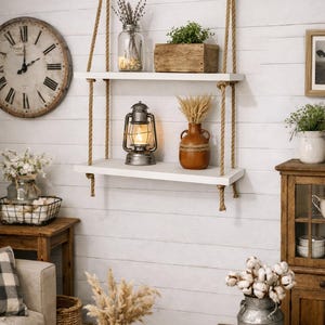May include: A two-tier wall-mounted shelf unit featuring two rectangular white wooden boards suspended by thick tan jute ropes. The ropes are secured at the top with metal rings, creating a hanging display for decorative items and home accessories.