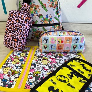 May include: A collection of bags and accessories with various patterns. Includes a gray backpack with Minecraft characters, a pink leopard print bag, a rectangular pencil case with cartoon dogs, a yellow mat with Hello Kitty characters, and a yellow Batman-themed accessory.