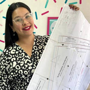 Puede incluir: Una mujer sostiene un patrón de costura grande. El patrón es blanco con texto y diagramas en negro. La mujer lleva una blusa con estampado en blanco y negro y gafas. La pared detrás tiene formas coloridas.