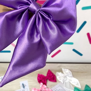 May include: A large purple satin bow with a pointed tip. The bow is made of a soft, silky fabric and has a smooth, shiny finish.