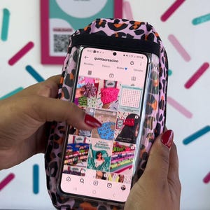 May include: A pink and black leopard print phone case with a clear front, displaying a phone screen. The phone screen shows an Instagram profile. The case is held by a person with red nail polish.