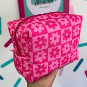 Puede incluir: Bolsa rosa con cremallera con un patrón a cuadros de flores rosas y blancas. La bolsa está hecha de tela y tiene un cierre de cremallera.